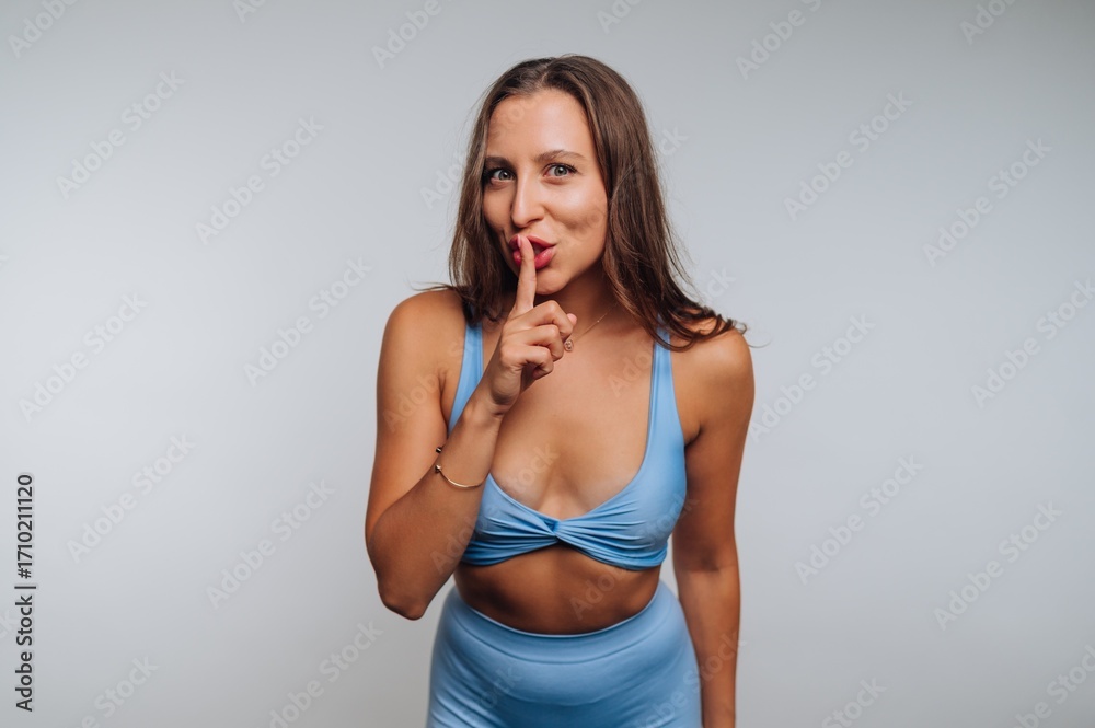 Fototapeta premium Woman in blue activewear holds a finger to her lips in a playful gesture, capturing attention in a simple studio setting