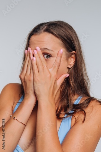 Woman covers her face with hands, showing a mix of surprise and shyness in a light blue outfit indoors
