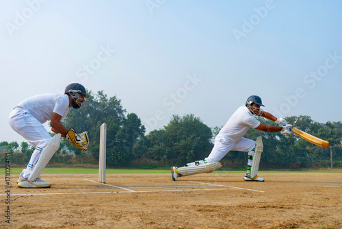 A batsman in white executes a powerful shot on a dirt field, with a wicketkeeper closely observing, highlighting teamwork and skill