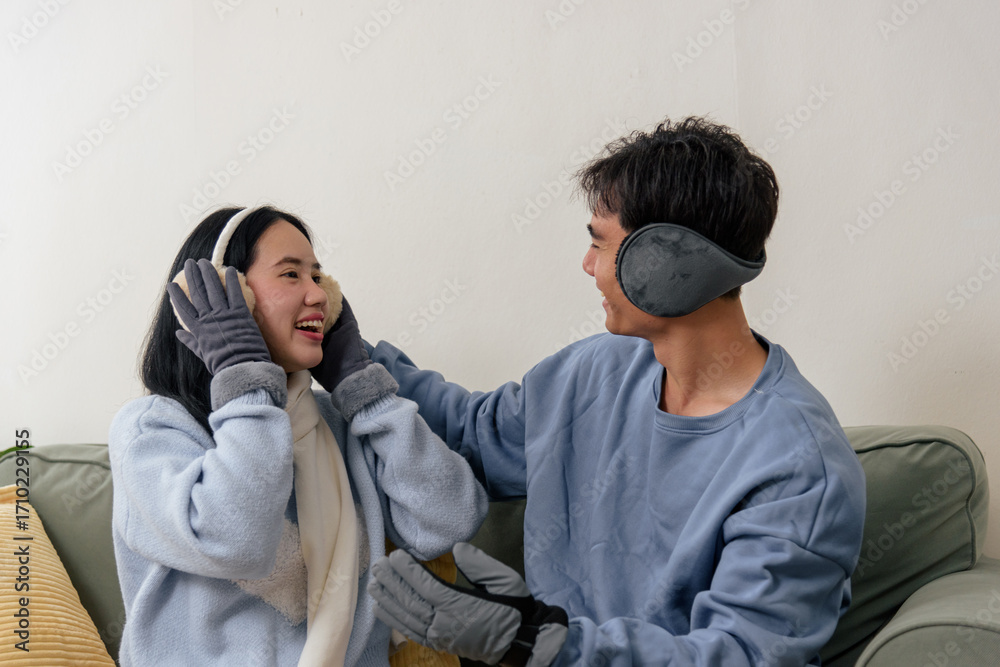 Fototapeta premium Winter cold day, happy couple, loving family inside home, people love each other, enjoying cozy time wearing warm winter clothes, smiling warmly on couch, earmuffs, and gloves