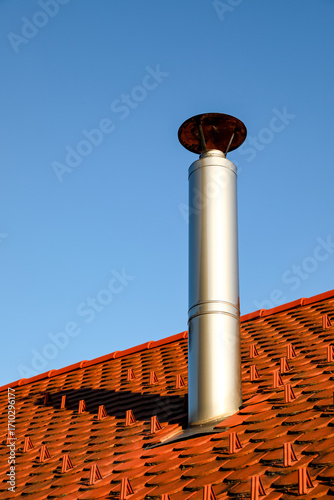 Polished metal chimney stands tall above a vibrant red tile roof under a clear blue sky. The round-capped flue gleams in sunlight, symbolizing efficient home heating and sleek architectural design.
