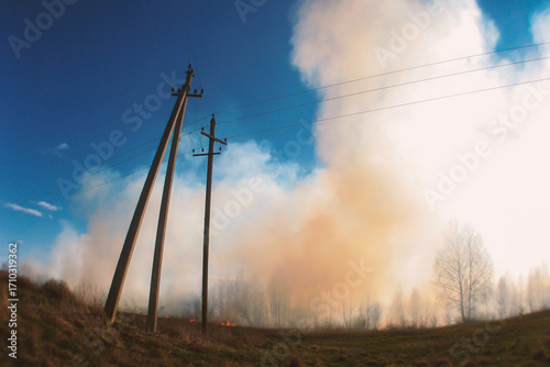 Rural field burning with thick smoke, under a blue sky, framed by strong utility poles.