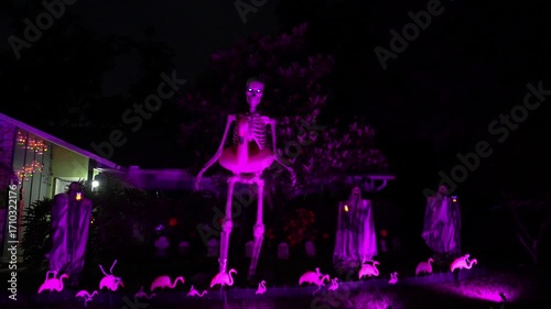 Front yard Halloween display with a giant skeleton, pink flamingos, ghosts, and tombstones glowing in purple light at night. Spooky suburban house scene shows neon lights, eerie decorations, and