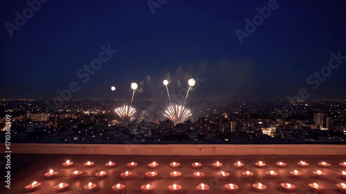Numerous candles arranged on a rooftop create a warm glow, contrasting with the vibrant bursts of fireworks illuminating the sprawling cityscape below