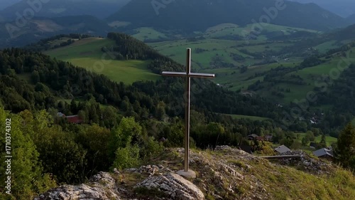 Cross on rocky hilltop overlooking green valley with forests and mountains in background