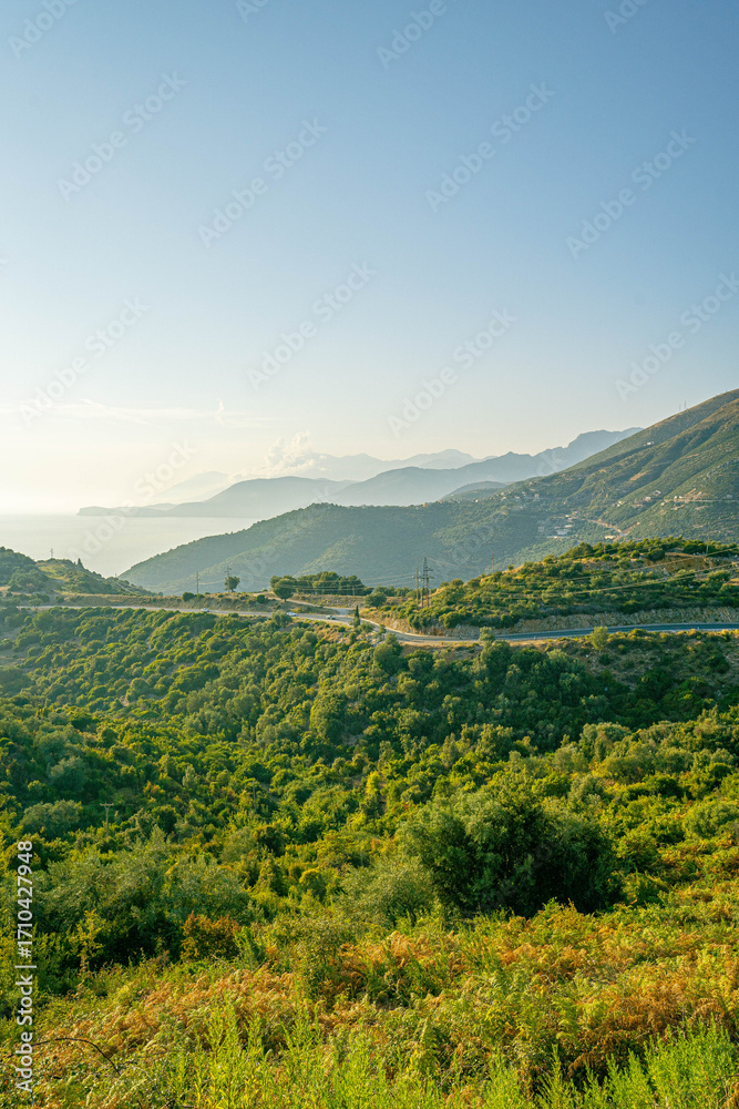 Fototapeta premium Scenic hillside with winding road leading towards sea and layered mountain ridges fading in horizon under bright blue sky in warm daylight