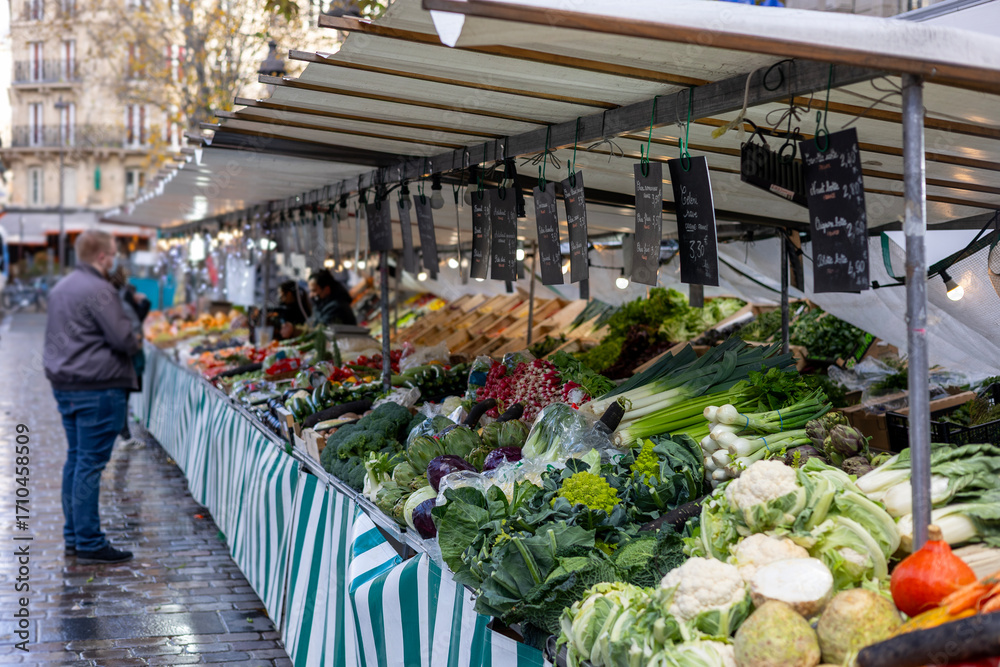 Fototapeta premium Fresh vegetables, including cauliflower, leeks, artichokes, radishes, kale, romanesco cabbage and cabbage, are for sale at a street market in Paris, France. A vendor is in the background.