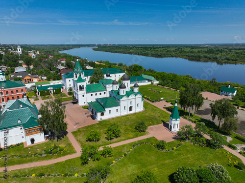 A wide aerial view of the Transfiguration Monastery with its white and green buildings and well-kept grounds, with the Oka River and bridge in the distance.