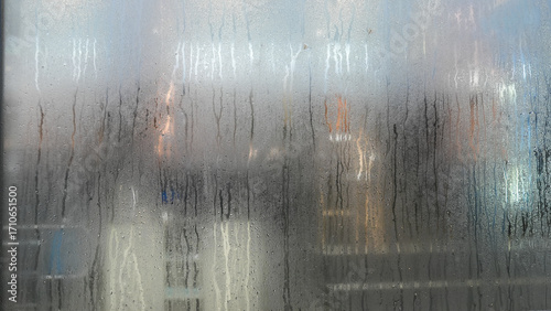 Close-up of wet glass with water droplets, distorting the view of warm glowing lights in the background.