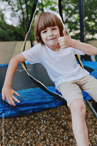 Valokuva Happy boy giving a thumb up while sitting on a trampoline, wearing a white shirt, enjoying a sunny day outdoors with a playful atmosphere