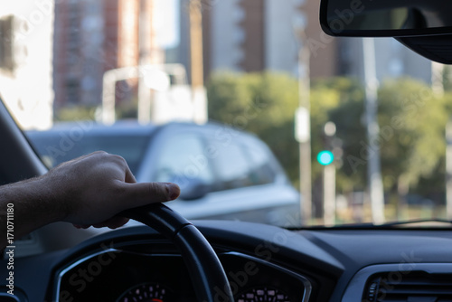 POV shot from inside a car, showing a hand on the steering wheel. The blurred cityscape and green traffic light in the background create a sense of movement and daily commute