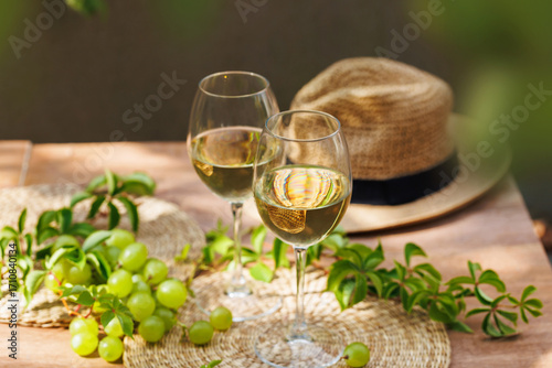 Two glasses of white wine with green grapes and straw hat on wooden patio table in natural light..