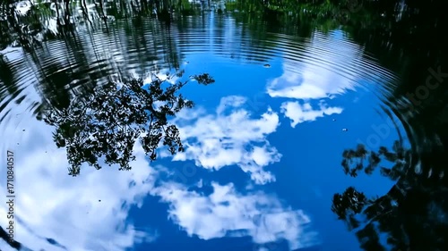 Serene ripples on a still pond reflect a vibrant blue sky filled with puffy clouds.