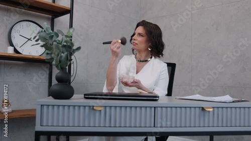 makeup applying, confident woman, desk makeup. In professional office setting, woman in white attire uses makeup brush to touch up her face at her workspace.