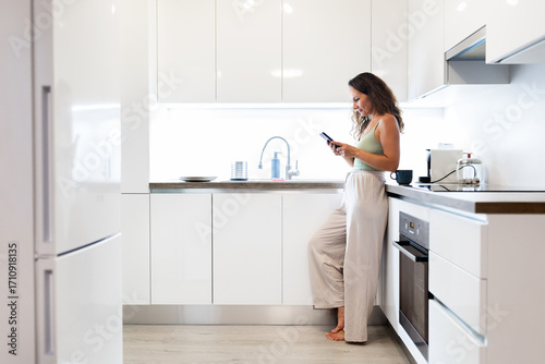 Cuadro en lienzo Young Caucasian woman at home kitchen leaning on counter while using smartphone