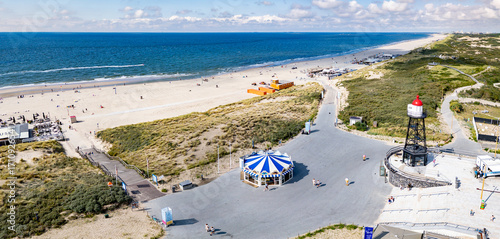 A panoramic view of a sandy beach of Kijkduin with a clear view of the sea. A lighthouse and a carousel are prominent near the shore, surrounded by pathways and greenery.