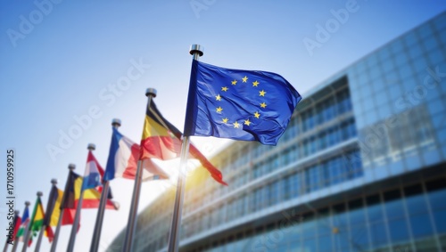 Low-angle shot of the European Union flag waving in bright sunlight with other European flags in front of a modern blurred office building
