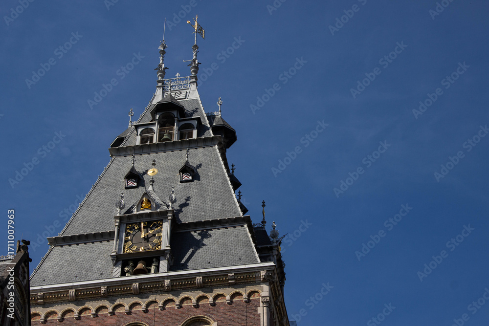 Fototapeta premium Architectural detail of a historic clock tower with intricate design elements, featuring a slate roof, decorative spires, and a clear blue sky backdrop, showcasing timeless craftsmanship