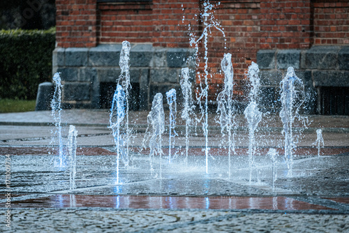 Jets of water shoot up from a ground fountain in front of a brick wall, lit by glowing blue lights, creating a dynamic and refreshing urban scene.