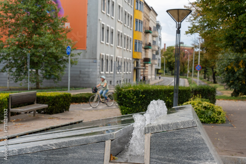 Modern urban fountain with water jets in city park with cyclist riding past colorful residential buildings and green landscaping. Concept of sustainable city living, bike-friendly infrastructure.