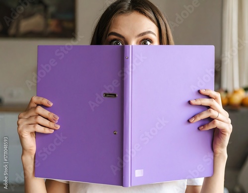 A woman holds a vibrant purple binder, concealing her face with its pages, conveying a playful or curious mood.