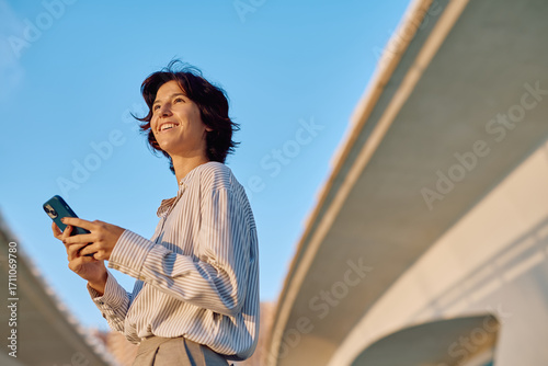 Fotografía Businesswoman smiling while using a smartphone under a bridge at sunset, enjoyin