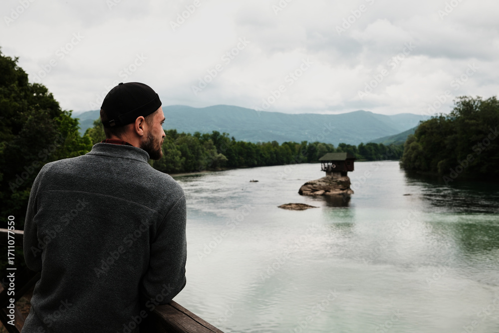 Fototapeta premium Man in a grey fleece jacket looking at the iconic house on the Drina River from a wooden viewpoint. Rear view. A popular tourist destination. Serbia country in spring season