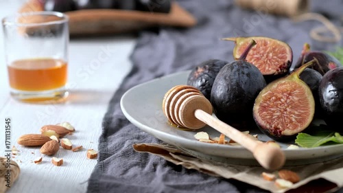 A rustic still life of fresh Black Mission figs, some whole and some cut in half to reveal their vibrant red flesh. The figs are arranged on a gray ceramic plate with a wooden honey dipper and chopped