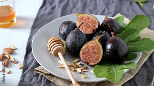 A rustic still life of fresh Black Mission figs, some whole and some cut in half to reveal their vibrant red flesh. The figs are arranged on a gray ceramic plate with a wooden honey dipper and chopped