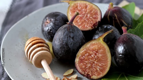 A rustic still life of fresh Black Mission figs, some whole and some cut in half to reveal their vibrant red flesh. The figs are arranged on a gray ceramic plate with a wooden honey dipper and chopped