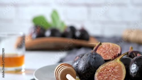 A rustic still life of fresh Black Mission figs, some whole and some cut in half to reveal their vibrant red flesh. The figs are arranged on a gray ceramic plate with a wooden honey dipper and chopped