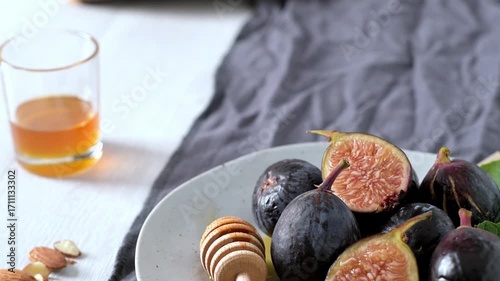 A rustic still life of fresh Black Mission figs, some whole and some cut in half to reveal their vibrant red flesh. The figs are arranged on a gray ceramic plate with a wooden honey dipper and chopped