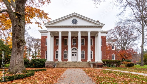 A two-story brick building featuring large white columns, surrounded by colorful autumn foliage and a pathway covered with fallen leaves