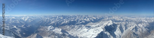 A wide aerial view showcases a vast, rugged mountain range covered in snow under a bright blue, cloudless sky