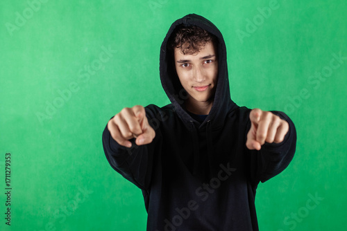 Portrait of a teenager in a black hoodie on a green background, pointing his hands forward