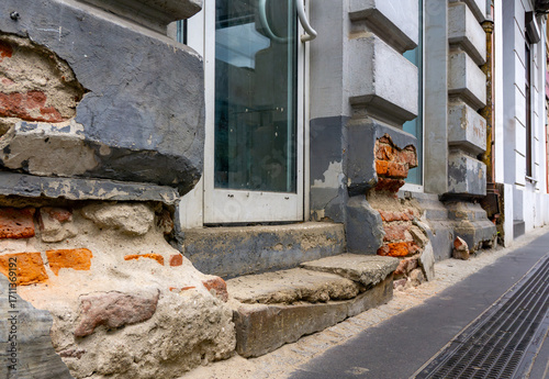 Fotografia Damaged tenement facade with exposed bricks and plaster loss, visible entrance with glass doors along urban sidewalk street