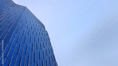 Quadro su tela Upward view of a sleek, reflective glass skyscraper with geometric patterns, rising against a bright, open blue sky in an urban setting
