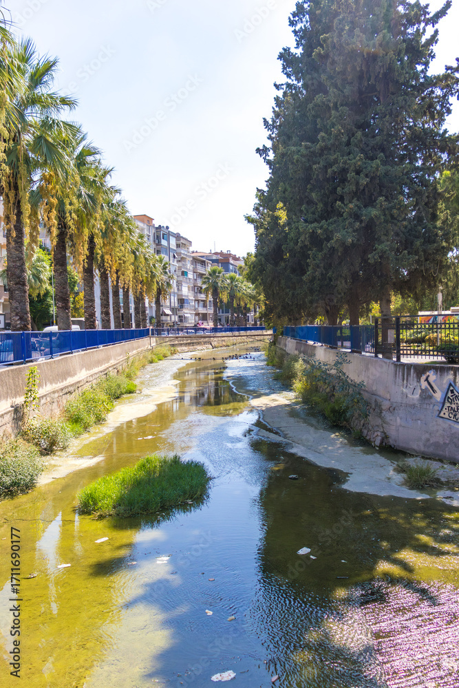 Fototapeta premium Urban river with dirty water, algae, and scattered litter in a modern city. Environmental pollution problem caused by urbanization and lack of waste management. Concept of climate change, water