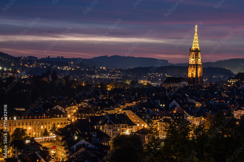 Fototapeta premium Bern, Switzerland, nighttime cityscape featuring illuminated buildings and a prominent tower, showcasing vibrant lights against a twilight sky, creating a serene urban atmosphere with captivating view