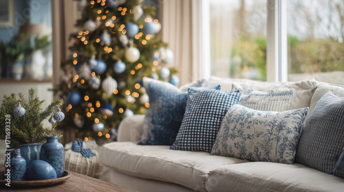 A cozy living room decorated for Christmas with blue and white ornaments, a glowing tree, and patterned pillows on a white sofa near a bright window.
