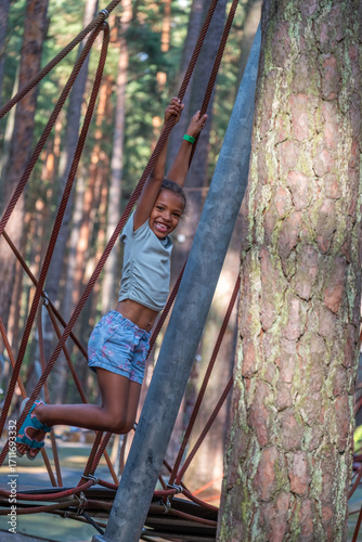 African American girl joyfully swinging on ropes in a forest playground, surrounded by tall trees, capturing the essence of childhood happiness and playful innocence