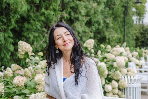 Elegant elder woman with long dark hair, wrapped in a soft shawl, enjoys a serene moment in a beautiful garden surrounded by blooming flowers, embodying wisdom and natural beauty