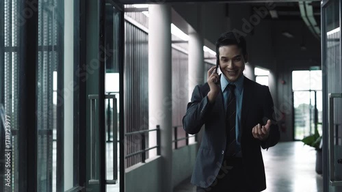 Wallpaper Mural Professional asian businessman walking through modern office corridor, talking on smartphone with confident smile and engaging hand gestures while navigating corporate interior Torontodigital.ca