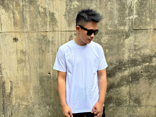 Portrait of young man wearing plain white short sleeve t-shirt and sunglasses standing against textured concrete wall