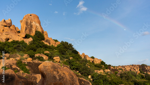 A view of Hampi's golden boulders with a rainbow and the moon at sunset.