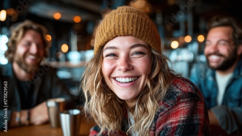 A cheerful young woman with curly hair wearing a beanie smiles brightly while sitting with friends in a cozy indoor setting filled with warm lights and a friendly atmosphere.