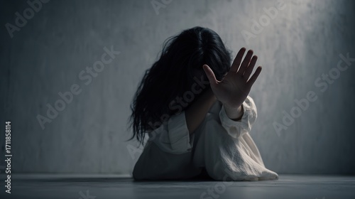 Sad woman with long black hair sitting on floor in white shirt, raising hand in defensive gesture, concept of stop violence against women, sexual abuse prevention and harassment awareness