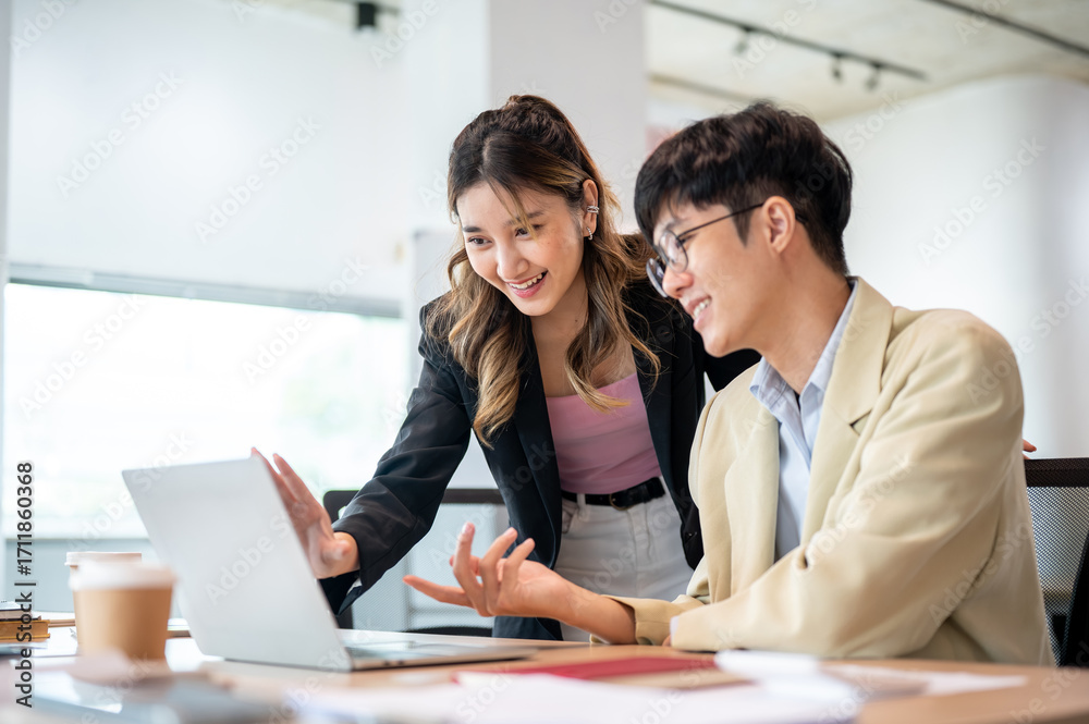 Fototapeta premium Smiling asian businesswoman looking at laptop talking with intern coworker at wooden table in office
