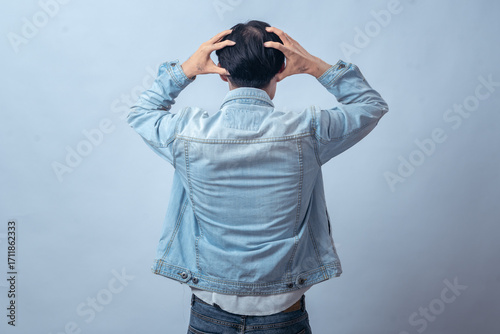 Asian man in denim jacket seen from behind, holding head with both hands in frustration, standing in studio against plain light background, expressing stress, tension, and emotional struggle