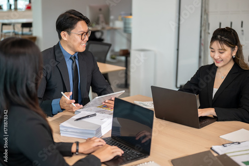 Asian businessman holding tablet talking to woman coworker while sitting at wooden table in office.